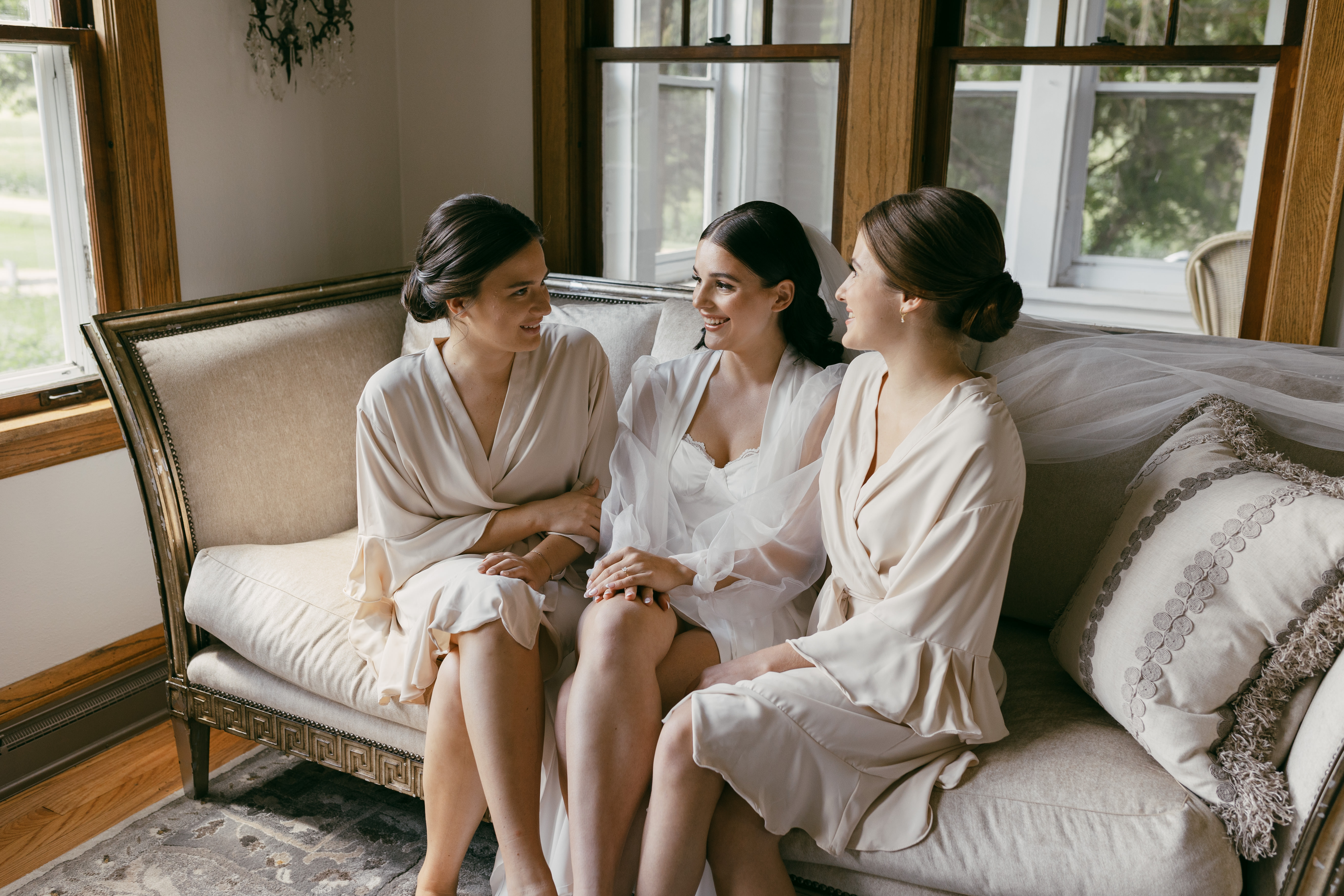 A bride sits closely with her sisters, who are serving as her bridesmaids, on a vintage sofa. The group is engaged in intimate conversation, talking and giggling together in a candid moment of sisterly bond during wedding day preparations. Their close positioning reflects the warmth and familiarity of their relationship.