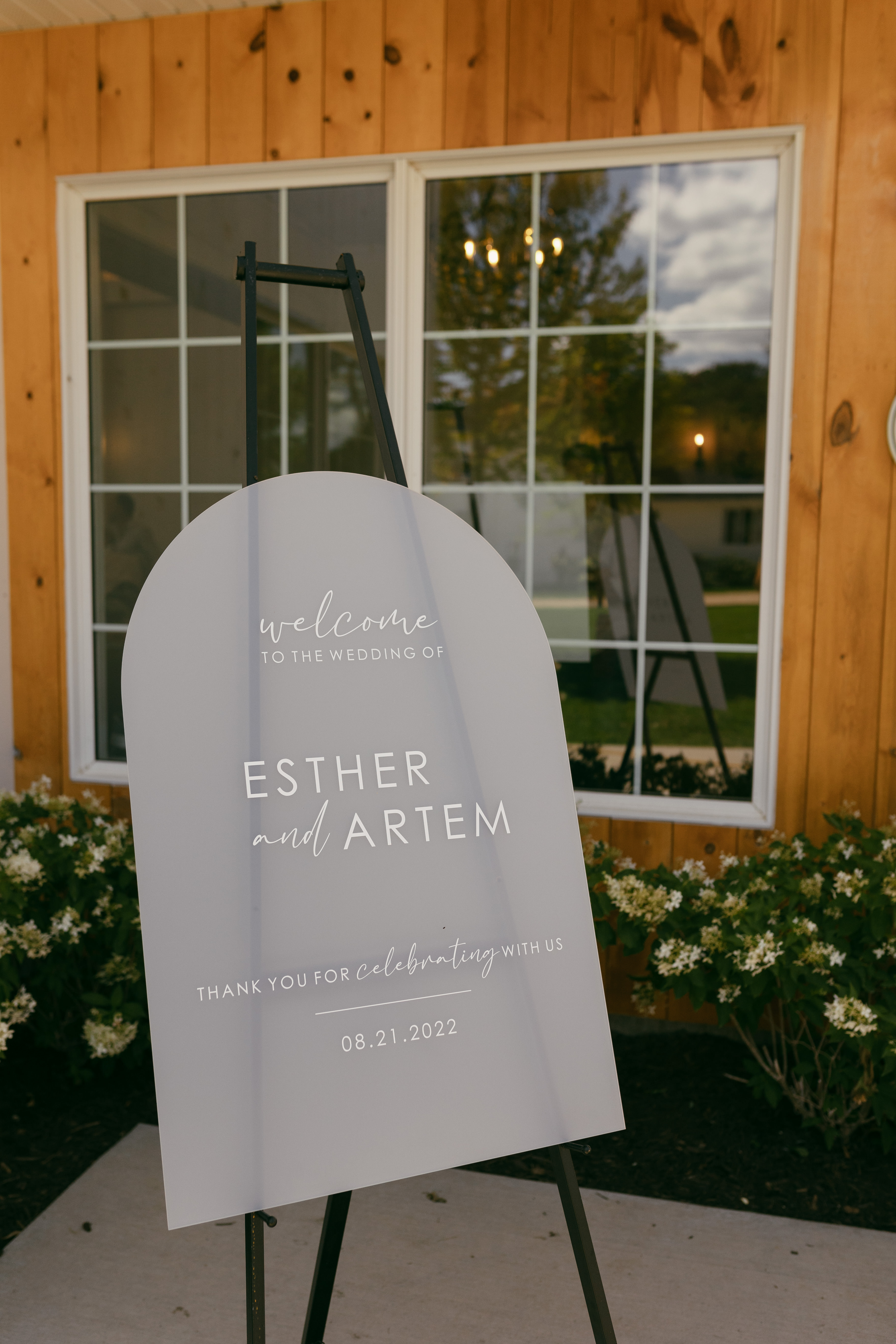 A semi-transparent welcome sign displayed on a wooden stand outside the couple's wedding reception space. The clear acrylic or glass sign reads 'Welcome' and serves as an elegant greeting for guests arriving at the celebration.