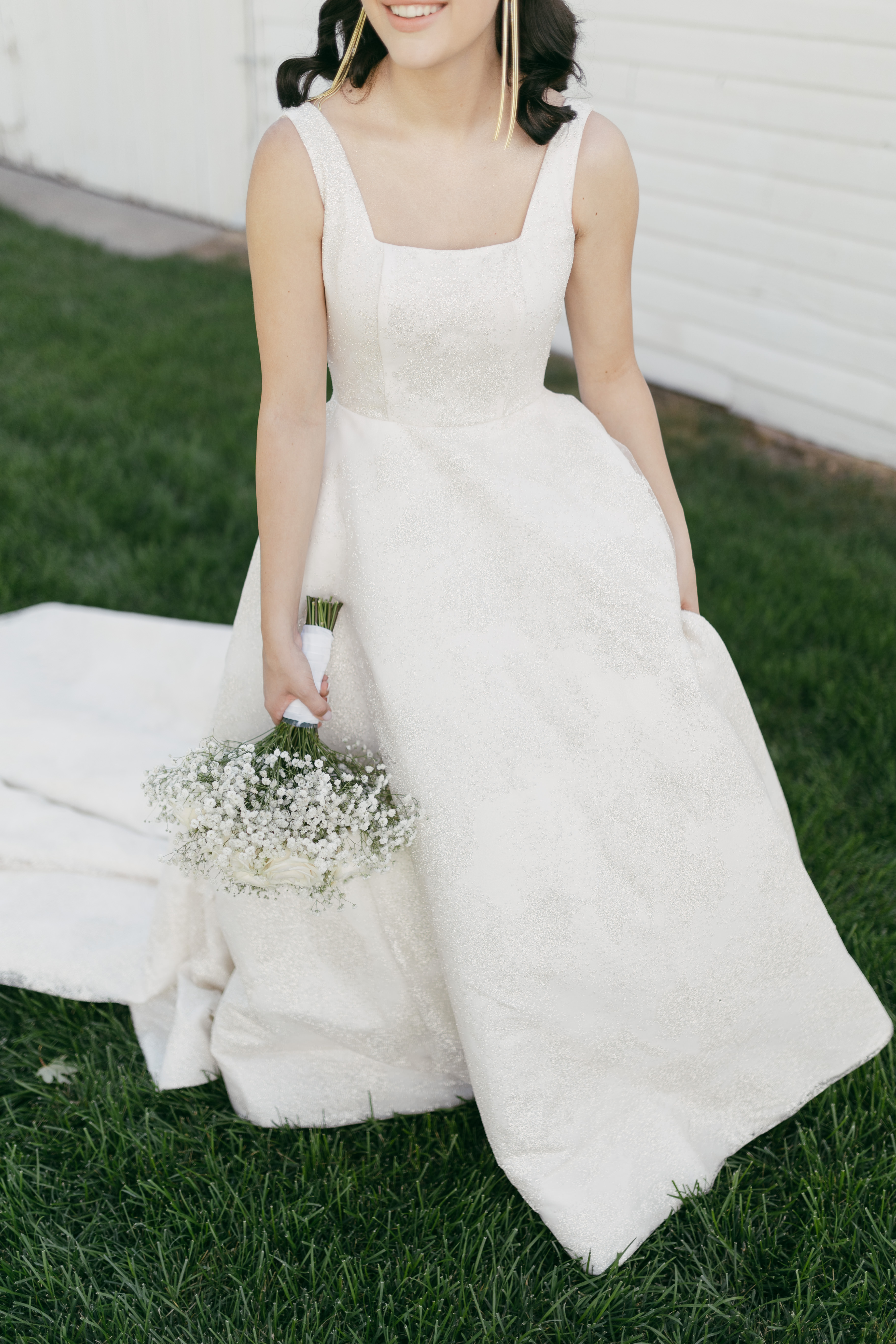 A detail photograph of a bride's wedding dress as she holds her bouquet at her side, taken outdoors. The image focuses on the dress fabric and silhouette without showing the bride's face, creating an elegant composition that highlights the gown's design alongside the bridal bouquet.