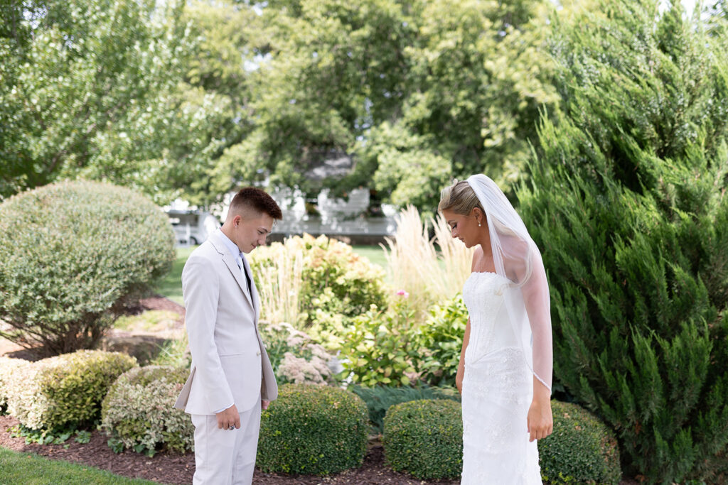 A bride in a white wedding gown and veil stands beside a groom in a light suit, both looking down in a garden setting with lush greenery in the background.
