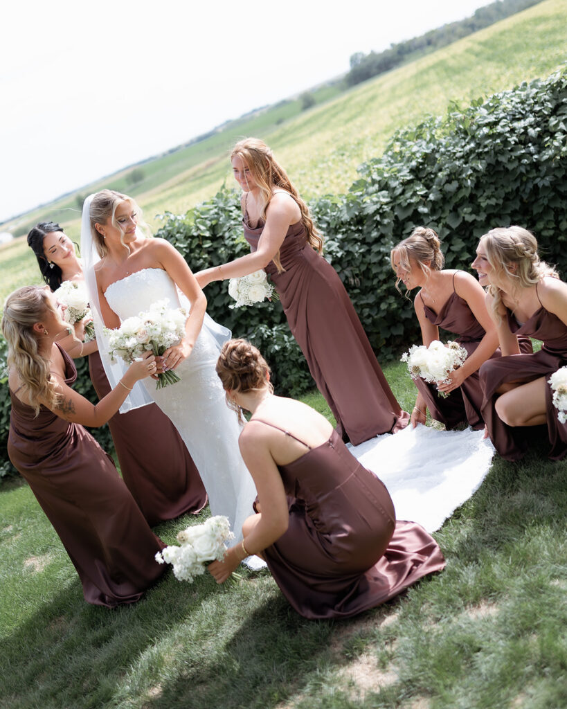 A bride in a strapless wedding gown surrounded by her bridesmaids in mocha satin dresses, adjusting her veil and dress in a lush outdoor setting.