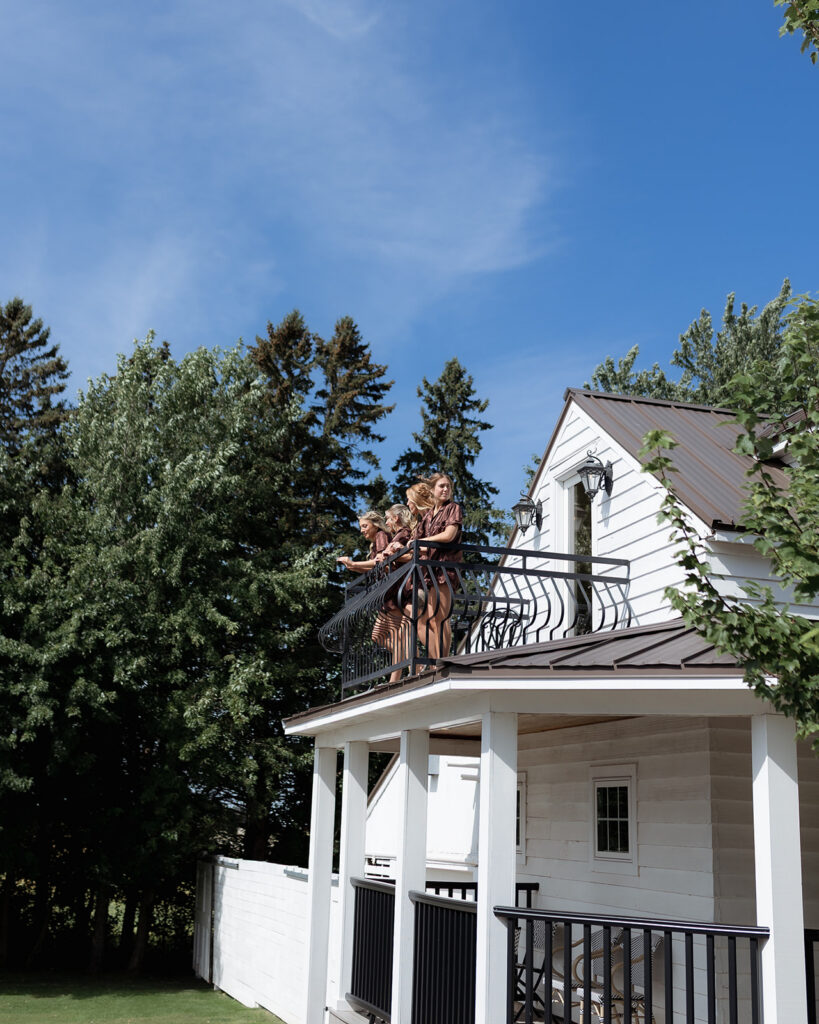 Four women in matching chocolate satin pajamas standing on a wrought iron balcony, overlooking a scenic view at Legacy Hill Farm, with green trees and a blue sky in the background.