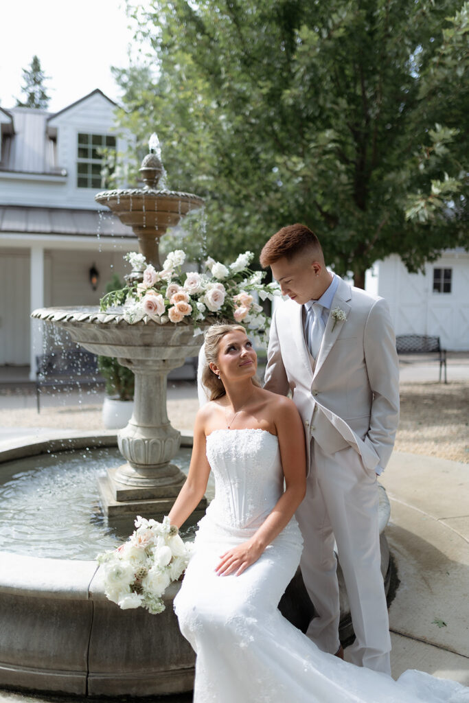 A bride in a strapless wedding gown sits by a tiered stone fountain decorated with flowers, gazing lovingly at her groom, who stands beside her in a light tan suit.