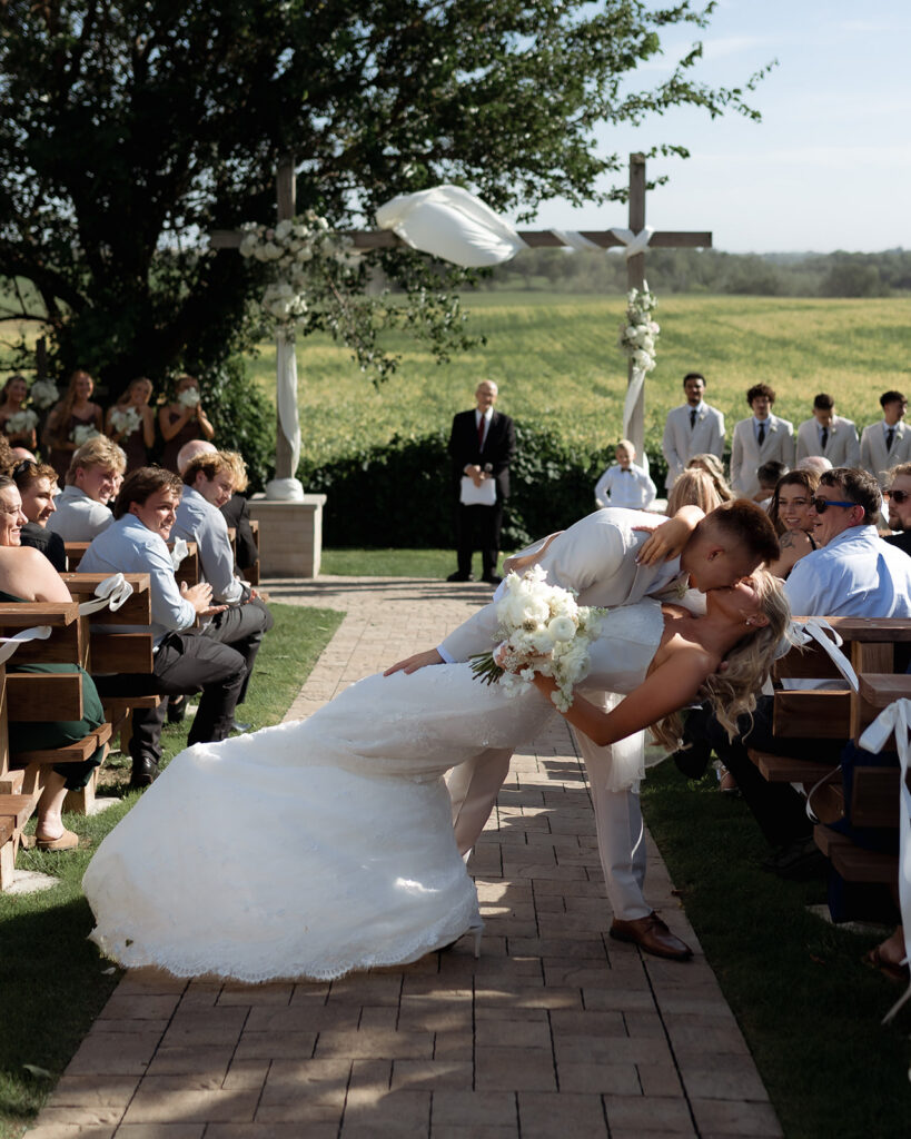 A bride and groom share a romantic kiss as the bride is dipped, surrounded by wedding guests at an outdoor ceremony with a scenic green backdrop.