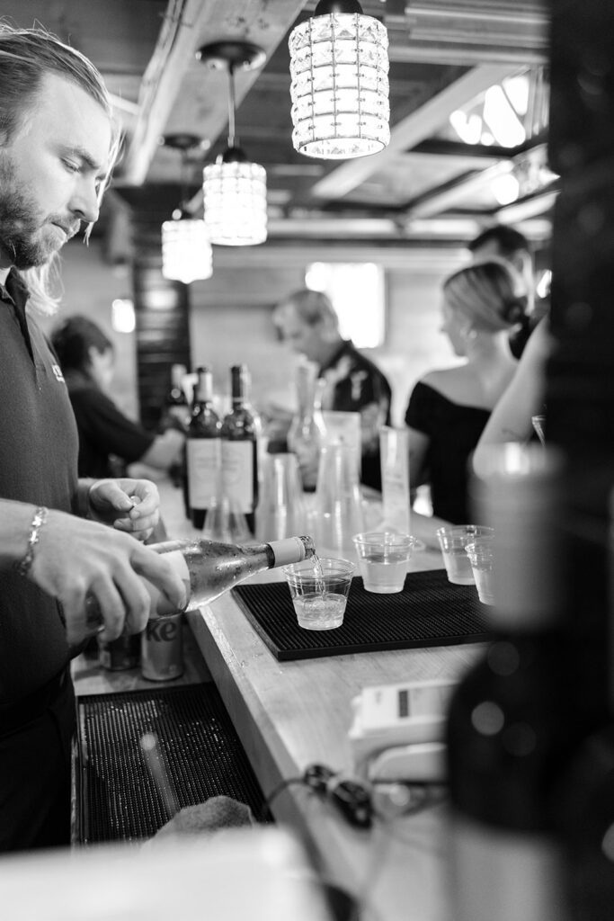 A bartender pouring drinks at a busy bar with dim lighting and hanging light fixtures.
