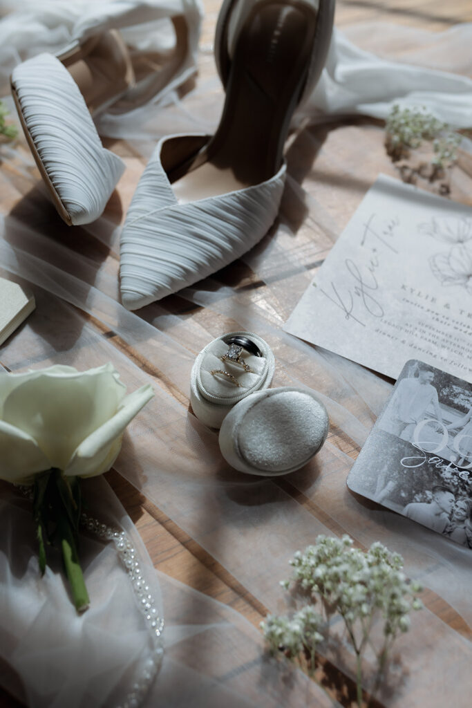 A flat lay of wedding details featuring pleated ivory heels, a velvet ring box with rings, an invitation suite with hand-drawn florals, matching vow books, a bottle of perfume, a white rose boutonniere, sprigs of baby's breath, and a pearl-edged veil on a wooden surface.