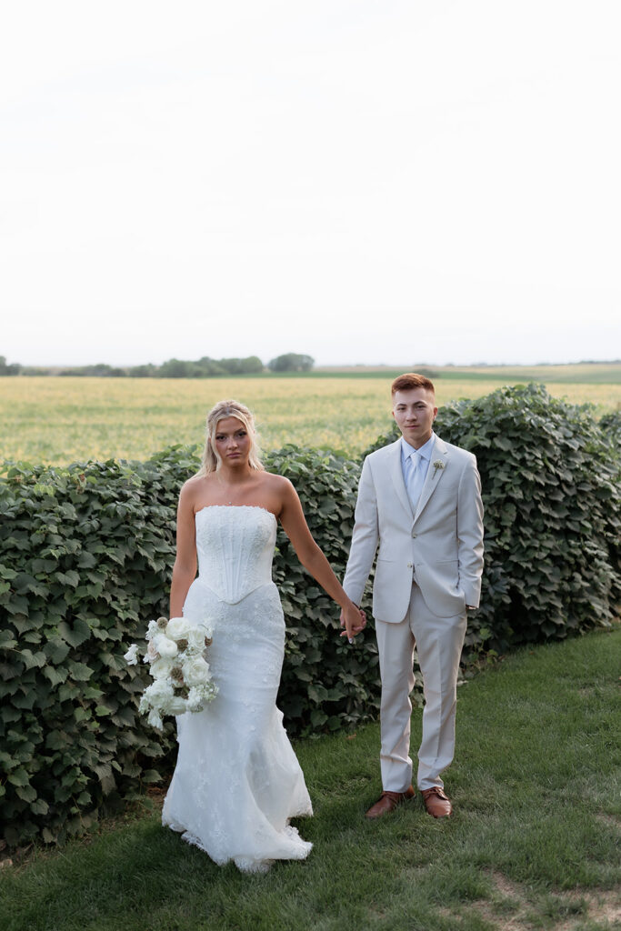 A couple stands hand in hand in a lush green setting, with the bride wearing a fitted strapless gown and holding a bouquet of white flowers, and the groom in a light tan suit, against a backdrop of rolling fields.
